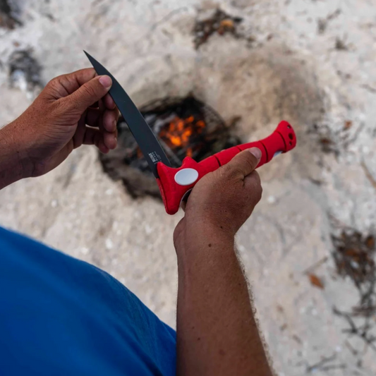 Person holding a knife with a red sheath near a campfire.