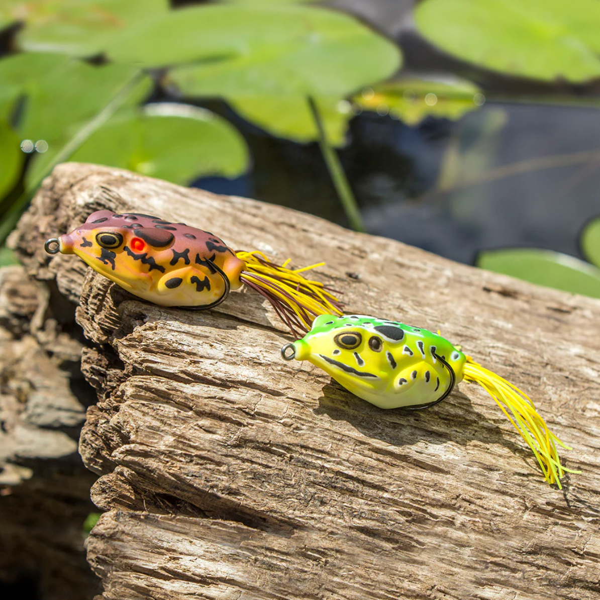 Two colorful frog-shaped fishing lures on a wooden log with water and lily pads in the background.