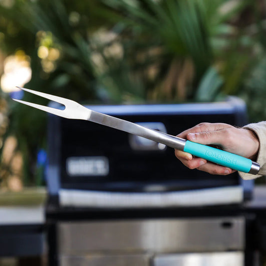 Person holding a grill fork with a turquoise handle in front of a barbecue grill.