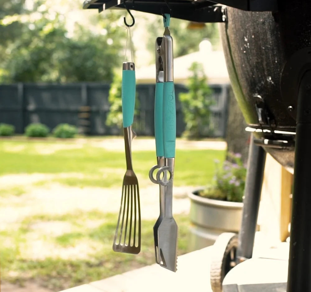 Teal-handled grill tools hanging from a vehicle's rearview mirror with a grassy area in the background.