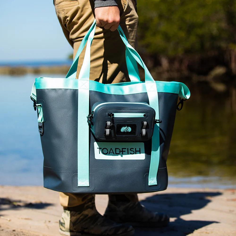 Person holding a teal and black tote bag with 'TOADFISH' branding by a body of water.