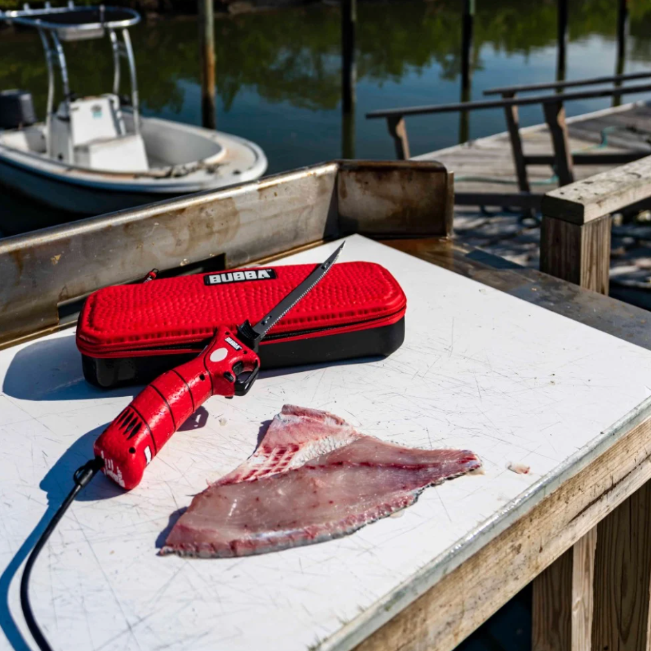 Red Bubba brand electric knife on a table with a fish next to it, dock setting in the background