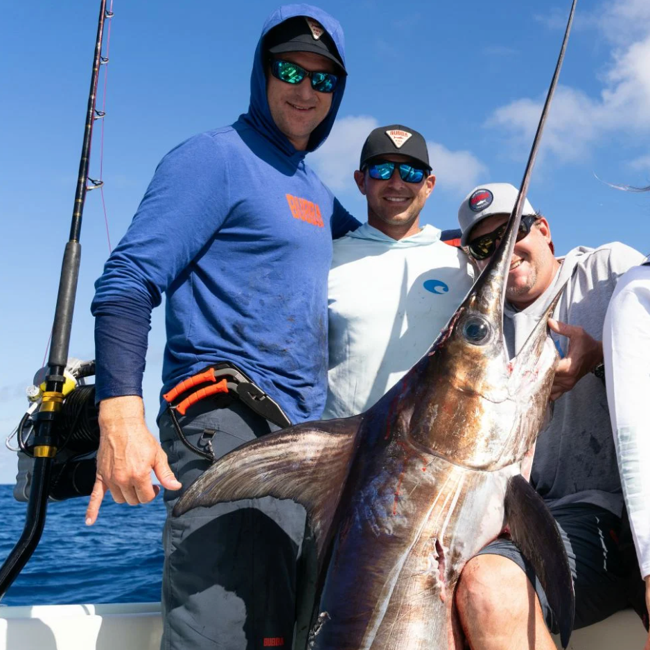 Three men on a boat holding a large fish with fishing equipment in the background.