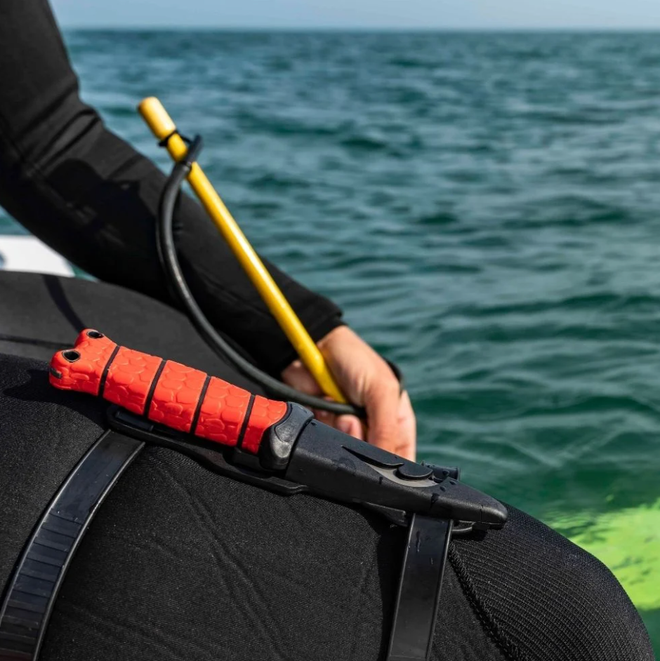 Person in wetsuit holding a red and black tool with ocean in background