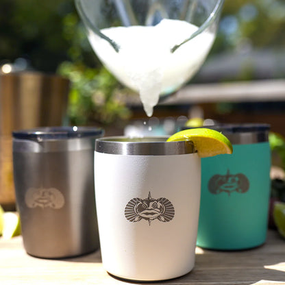 White insulated cup with a logo, surrounded by other insulated cups and a glass with ice, on a wooden surface outdoors.