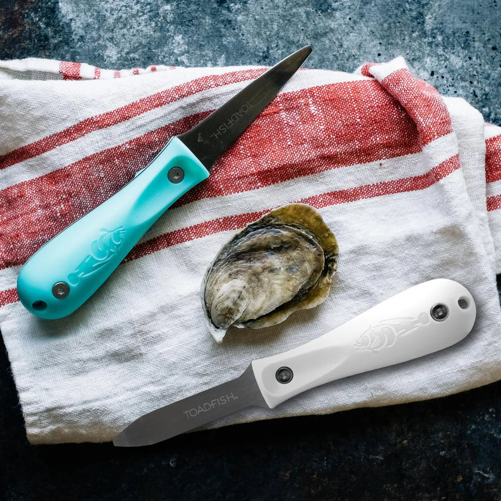 Two oyster shucking knives with turquoise and white handles on a red and white striped towel with an oyster shell.