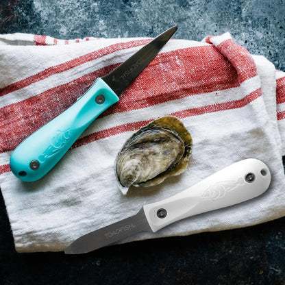 Two oyster shucking knives with turquoise and white handles on a red and white striped towel with an oyster shell.