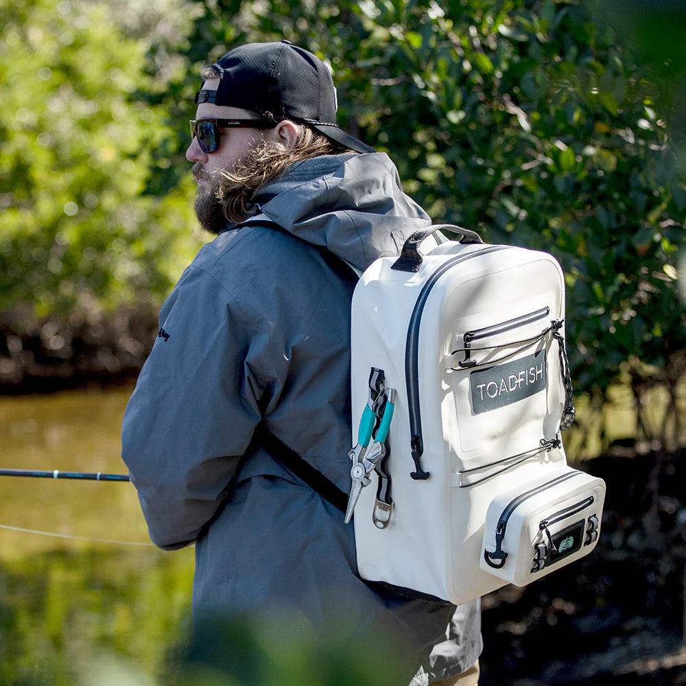 Man with a white backpack labeled 'TOADFISH' standing by a body of water with greenery.