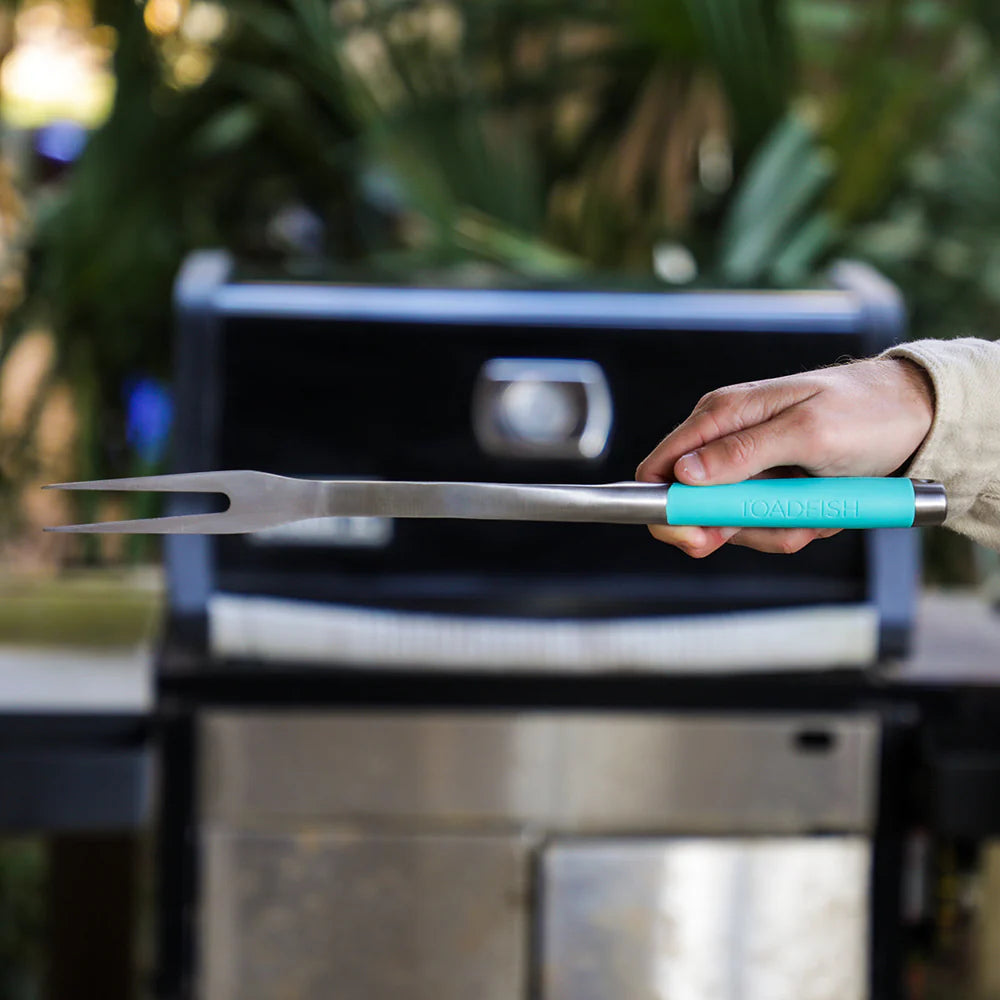 Person holding a grill fork with a turquoise handle in front of a barbecue grill.