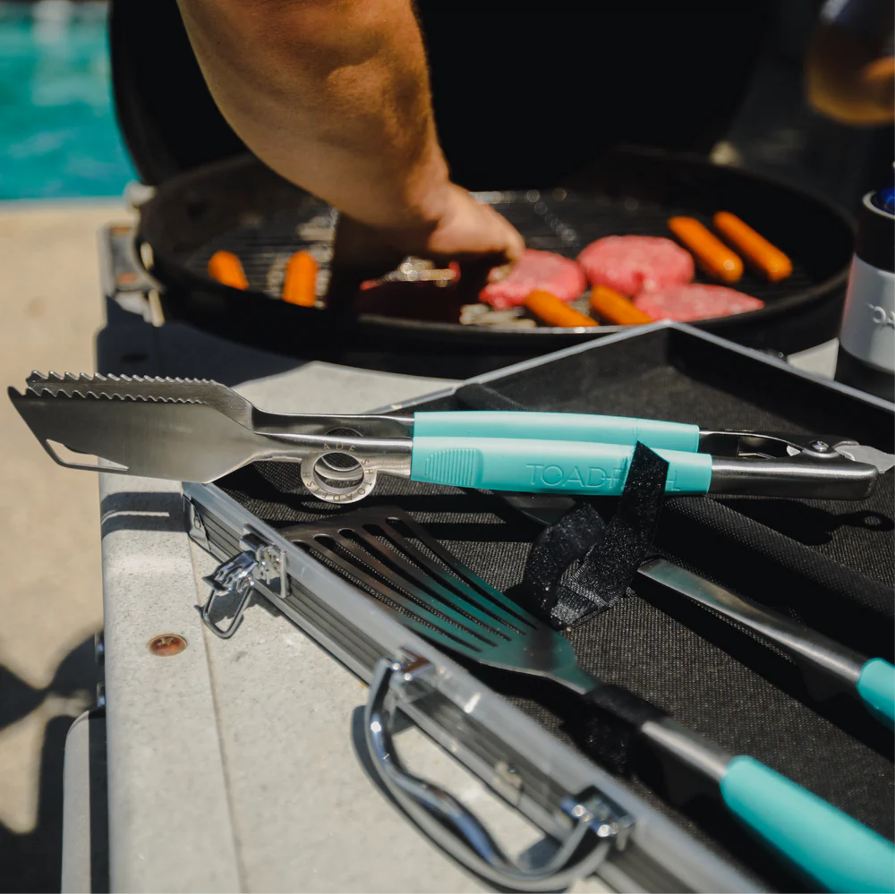 Barbecue tools with turquoise handles on a grill next to a pan of food.