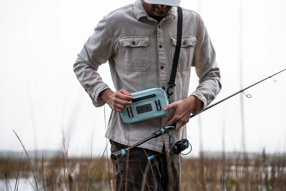 Man holding a teal cooler box by a lake with fishing equipment.