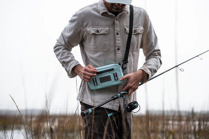 Man holding a teal cooler box by a lake with fishing equipment.