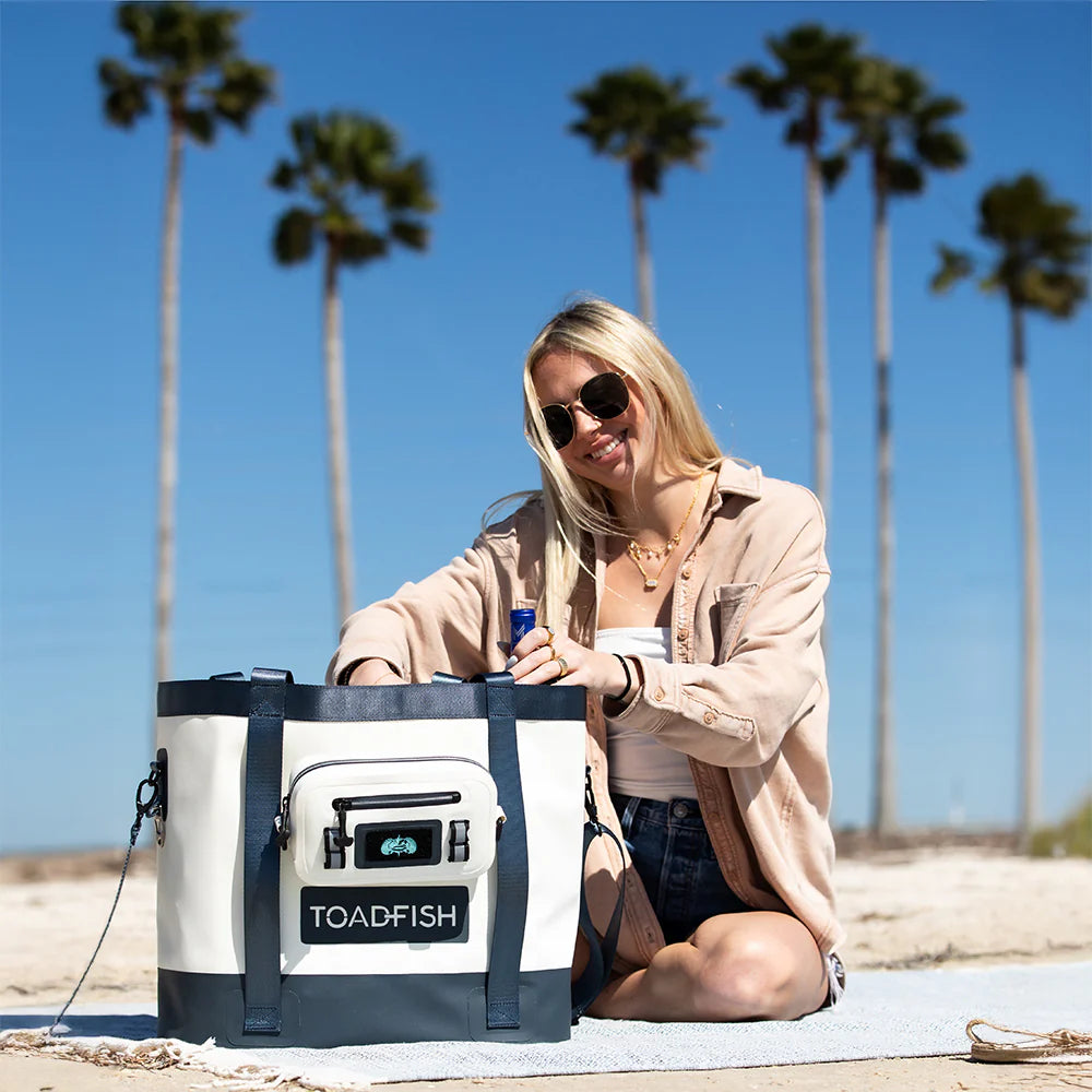 Woman sitting on a beach with a large cooler bag branded 'TOADFISH' in the foreground.